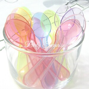 May include: A clear glass bowl filled with colorful, translucent plastic spoons. The spoons are in shades of pink, orange, yellow, green, and purple. The text "Party Surprise" is visible on the side of the bowl.