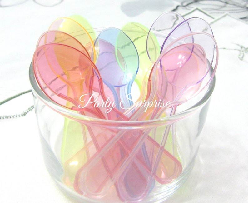 May include: A clear glass bowl filled with colorful, translucent plastic spoons. The spoons are in shades of pink, orange, yellow, green, and purple. The text "Party Surprise" is visible on the side of the bowl.