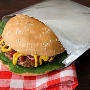 May include: A hamburger with a sesame seed bun, lettuce, ketchup, and mustard is wrapped in aluminum foil and placed on a red and white checkered picnic blanket.