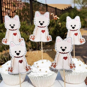 May include: Five white cupcake toppers with a cartoon image of a white dog with a red heart on its chest. The toppers are on wooden sticks and are inserted into white cupcakes with gold sprinkles.