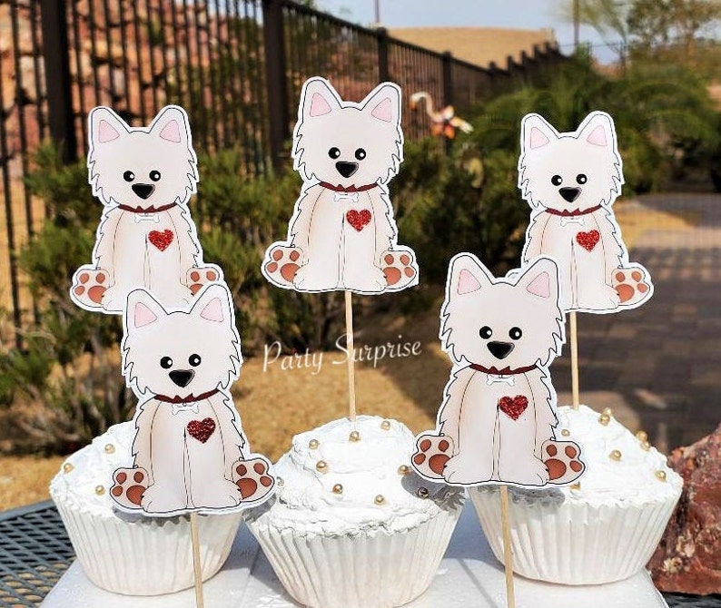 May include: Five white cupcake toppers with a cartoon image of a white dog with a red heart on its chest. The toppers are on wooden sticks and are inserted into white cupcakes with gold sprinkles.