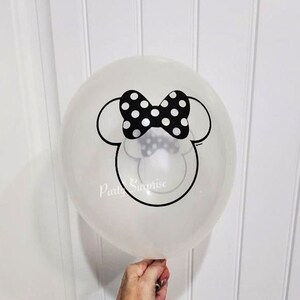 May include: A clear balloon featuring a black and white Minnie Mouse head design. The design includes a black bow with white polka dots and Minnie's signature ears.