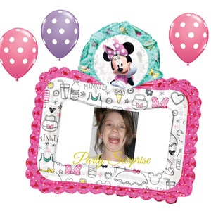 May include: A Minnie Mouse-themed party decoration set. Includes a pink and white polka dot frame with a photo, three balloons, and a Minnie Mouse-shaped balloon. The frame has a scalloped edge and is decorated with Minnie Mouse-themed graphics.