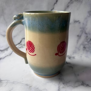 May include: A handmade ceramic mug with a beige body and a blue and green rim. Two red yarn ball designs are printed on the mug. The handle is beige with a small decorative element at the top.