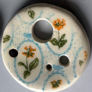 May include: A round, white ceramic button featuring a repeating pattern of orange flowers with green leaves enclosed in light blue oval frames. The button has four circular holes of varying sizes distributed across its surface.
