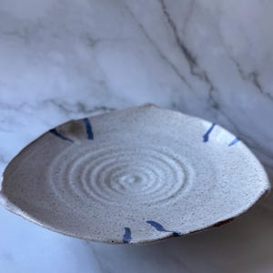 May include: A white ceramic serving dish with a speckled glaze and blue painted lines. The dish has a slightly irregular shape and a textured surface.