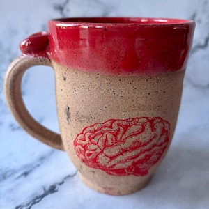 May include: A handmade ceramic mug with a red interior and rim. The exterior is tan with a red brain graphic. The handle has a small red accent. This unique mug is perfect for coffee, tea, or any beverage.
