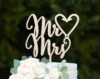 Peut inclure: Un gâteau de mariage blanc à deux étages avec un topper en bois "Mr & Mrs" et des fleurs blanches. Le gâteau est sur un support à gâteau vert menthe.