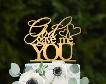 Puede incluir: Un pastel blanco de dos pisos con un adorno dorado que dice "God gave me YOU". El pastel está decorado con flores blancas y vegetación, y se encuentra sobre un soporte para pasteles verde menta. El fondo es verde borroso.