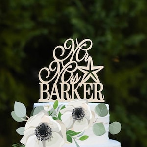 Könnte beinhalten: Eine zweistöckige weiße Hochzeitstorte, verziert mit weißen Anemonen und Grün. Die Torte ist mit einem hölzernen Tortendekor verziert, auf dem "Mr & Mrs Barker" mit einem Stern steht.