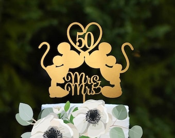 May include: A white two-tiered cake decorated with white flowers and greenery. A gold Mickey Mouse and Minnie Mouse cake topper with a heart that says "50" is on top of the cake. The cake is on a mint green cake stand.