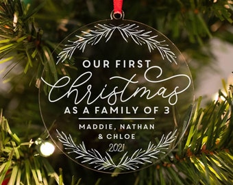 May include: Clear, round Christmas ornament with the text "OUR FIRST Christmas AS A FAMILY OF 3" in white script. The names Maddie, Nathan & Chloe, and the year 2021 are also included. The ornament hangs from a red ribbon on a Christmas tree.