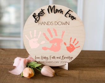 May include: A round wooden sign with the text "Best Mom Ever Hands Down" and three pink handprints. The sign is on a stand and has the text "Love, Hailey, Faith and Brooklyn Mother's Day 2024" at the bottom.