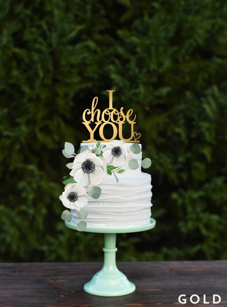 Puede incluir: Un pastel de boda blanco de dos niveles decorado con an&eacute;monas blancas y vegetaci&oacute;n. El adorno de la torta es dorado y dice "I choose YOU".
