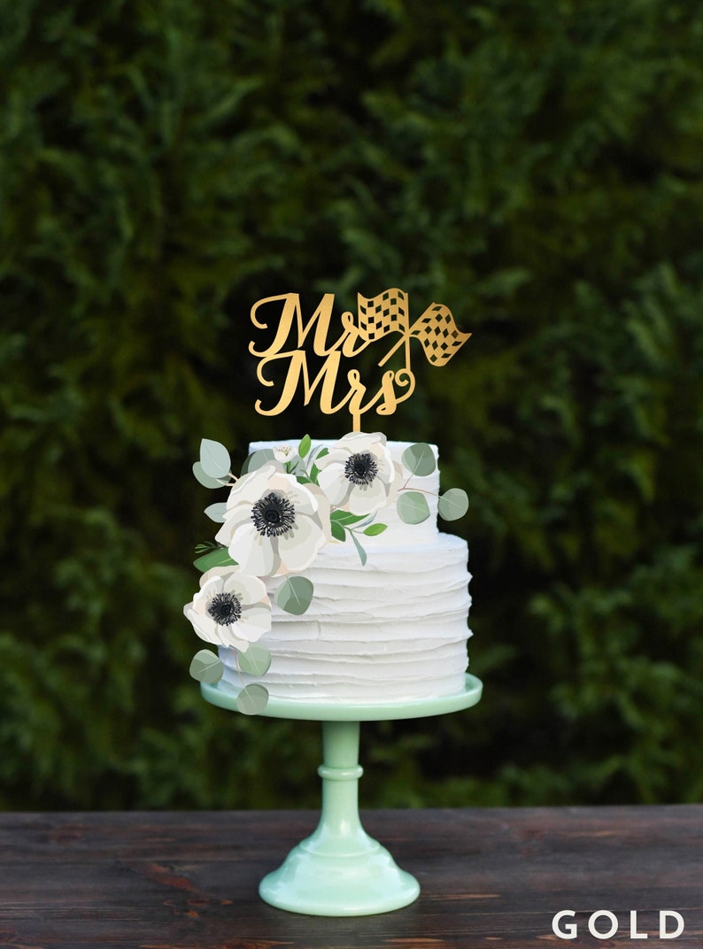 K&ouml;nnte beinhalten: Eine zweist&ouml;ckige wei&szlig;e Hochzeitstorte mit einem goldenen "Mr. & Mrs." Topper, der eine karierte Flagge zeigt. Die Torte ist mit wei&szlig;en Blumen und Gr&uuml;n verziert.