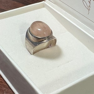 May include: A silver ring with a large, oval-shaped, light pink gemstone. The ring is in a white box with a white background.