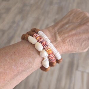 May include: Three stacked bracelets. One bracelet features large, off-white, oval-shaped beads. Another has a mix of orange, pink, and white shell beads. The third bracelet has white beads. All bracelets have brown wooden beads.