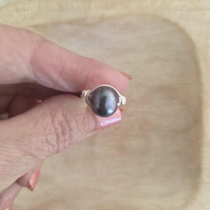 May include: A silver wire-wrapped ring featuring a dark gray, lustrous pearl. The ring's band is intricately coiled, showcasing the pearl's smooth, oval shape. The ring is held against a light wooden surface.