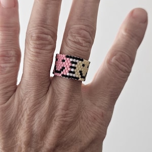 May include: A beaded ring in pink, black, and cream colors. The ring is worn on a finger and features a geometric pattern. The ring is approximately 1 inch wide.