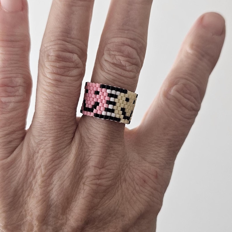 May include: A beaded ring in pink, black, and cream colors. The ring is worn on a finger and features a geometric pattern. The ring is approximately 1 inch wide.