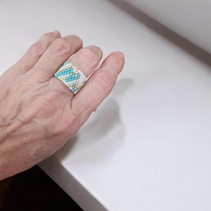 May include: A wide beaded ring in shades of turquoise, gold, and white. The ring features a geometric pattern and is worn on a finger. The ring is made of small, colorful beads, creating a textured surface. The ring is a fashion accessory.