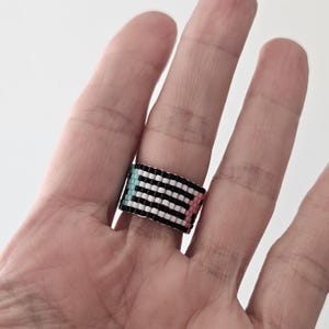 May include: A beaded ring with a geometric pattern. The ring features alternating black and white stripes, with sections of light blue and pink. The ring is worn on a finger.