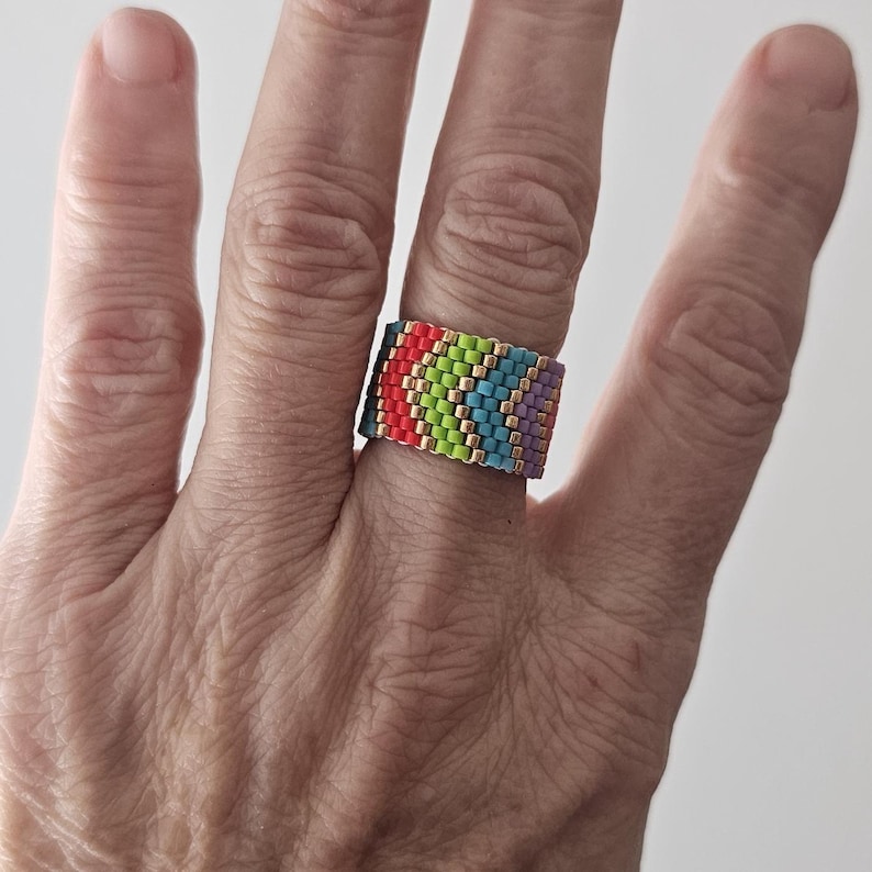 May include: A colorful beaded ring with a rainbow pattern of red, green, blue, and purple beads. The ring is worn on a finger and features gold-colored accents between the rows of beads. The ring is a unique piece of jewelry.