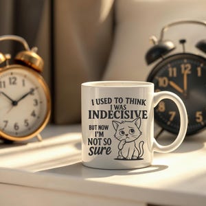 May include: White ceramic coffee mug with a black and white illustration of a cat. The mug has text that reads "I used to think I was indecisive but now I'm not so sure".