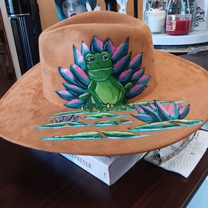 May include: A brown suede cowboy hat with a hand-painted design of a green frog sitting on lily pads. The frog is surrounded by pink and blue water plants. The hat is resting on a white book.