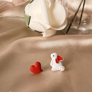 Può includere: Una statuetta di coniglio bianco con orecchie rosse e un accento di perla, e una statuetta di cuore rosso su uno sfondo di tessuto beige.