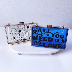 Può includere: Due pochette rigide in acrilico trasparente con dettagli dorati. Una pochette presenta una grafica in bianco e nero di cattivi Disney con il testo "Bad Girls Have More Fun". L'altra pochette è blu con il testo "All You Need Is Love" in nero.