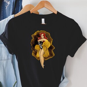 Pu&ograve; includere: Una maglietta nera con una grafica di una donna dai capelli rossi con un vestito giallo e guanti neri. La donna &egrave; in piedi in una luce dorata.