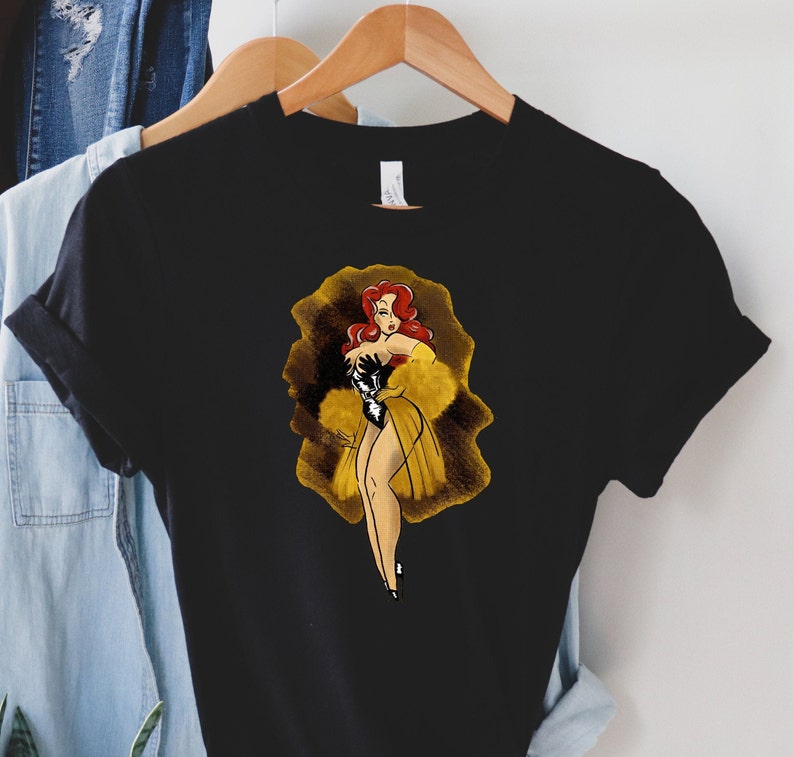 Pu&ograve; includere: Una maglietta nera con una grafica di una donna dai capelli rossi con un vestito giallo e guanti neri. La donna &egrave; in piedi in una luce dorata.
