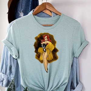 Pu&ograve; includere: Una t-shirt blu chiaro con un'illustrazione in stile vintage di una donna con un vestito giallo e capelli rossi. La donna tiene un boa di piume nere e si trova su uno sfondo dorato.