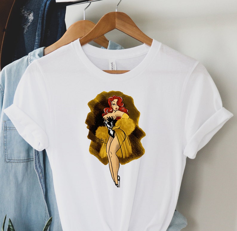 Pu&ograve; includere: Maglietta bianca con un'illustrazione di cartone animato di una donna con i capelli rossi che indossa un abito nero e giallo. La donna &egrave; in piedi su uno sfondo dorato e nero.