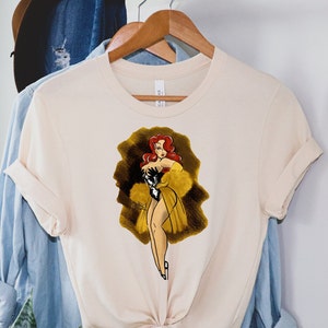 Pu&ograve; includere: Una t-shirt color crema con un'illustrazione in stile vintage di una donna con i capelli rossi che indossa un abito bianco e nero e un cappotto di pelliccia giallo. L'illustrazione &egrave; su uno sfondo marrone.
