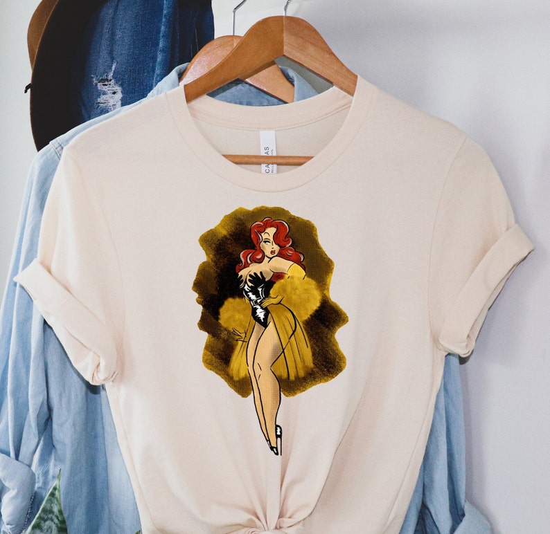 Pu&ograve; includere: Una t-shirt color crema con un'illustrazione in stile vintage di una donna con i capelli rossi che indossa un abito bianco e nero e un cappotto di pelliccia giallo. L'illustrazione &egrave; su uno sfondo marrone.