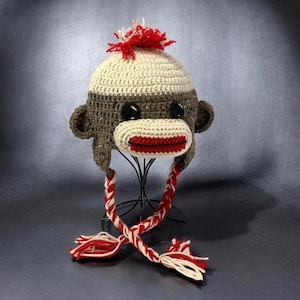 May include: A handmade crochet monkey hat in gray, cream, and red. The hat features a monkey face with black eyes, a red mouth, and ear flaps. The hat has a red and white braided tie and a red and white pom-pom on top.