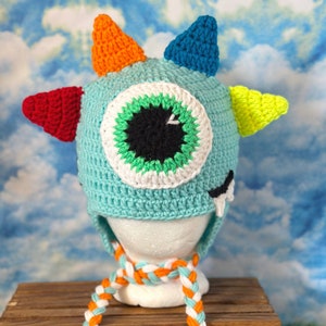 May include: A blue crocheted monster hat with a large green eye, orange, red, blue, and yellow horns, and a white and orange braided scarf.