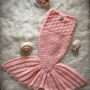 May include: A pink crocheted mermaid tail costume with a ruffled tail fin. The tail is decorated with a small, sparkly button.