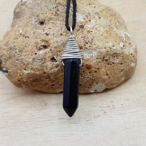May include: Black gemstone pendant with silver wire wrapping, hanging from a black cord. The pendant is shaped like a point and is resting on a light brown rock.