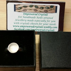 May include: A silver ring with a white stone set in a bezel setting. The ring is in a black box with a white card that says "Empowered Crystals" and has contact information for the business.