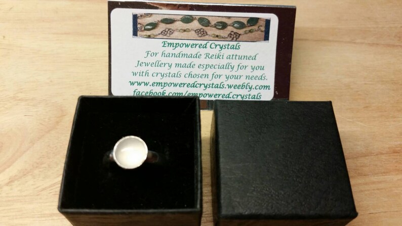 May include: A silver ring with a white stone set in a bezel setting. The ring is in a black box with a white card that says "Empowered Crystals" and has contact information for the business.