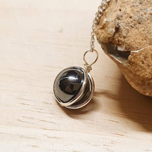 May include: A silver necklace featuring a polished, dark gray, spherical pendant. The pendant is encased in a silver wire cage and hangs from a delicate silver chain. The necklace is displayed on a light wooden surface, with a textured rock in the background.