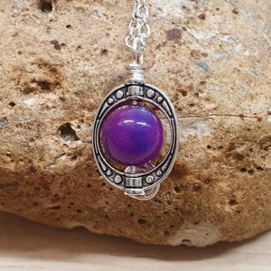 K&ouml;nnte beinhalten: Eine silberne Halskette mit einem violetten Glaskugelpendant in einem dekorativen Rahmen. Der Anh&auml;nger h&auml;ngt an einer silbernen Kette.