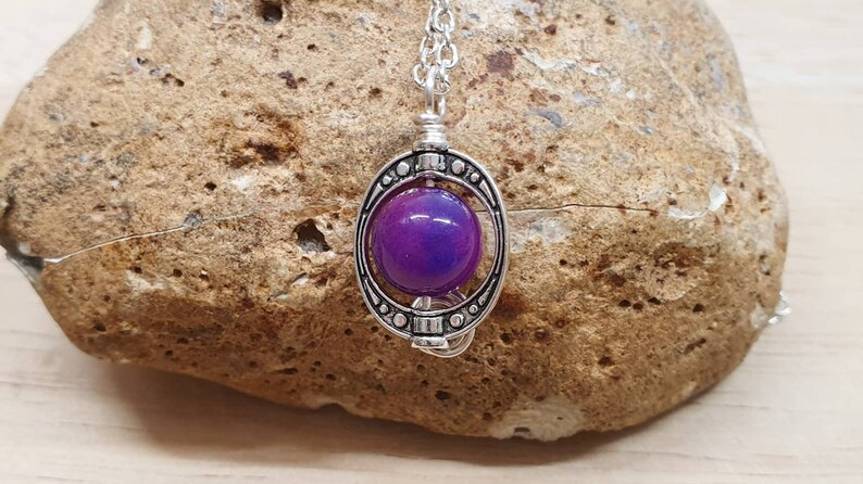 K&ouml;nnte beinhalten: Eine silberne Halskette mit einem violetten Glaskugelpendant in einem dekorativen Rahmen. Der Anh&auml;nger h&auml;ngt an einer silbernen Kette.