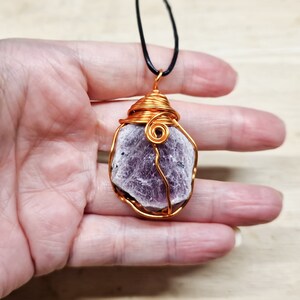 以下が含まれることがあります： 薄いピンクと紫の宝石を特徴とする、銅線で巻かれたペンダント。ペンダントは黒いコードからぶら下がっています。