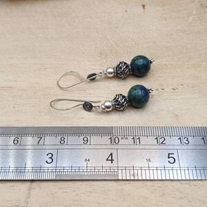 May include: A pair of handcrafted earrings featuring dark blue and green stone beads, silver-colored metal beads, and silver-colored ear wires. The earrings are displayed next to a ruler for size reference.