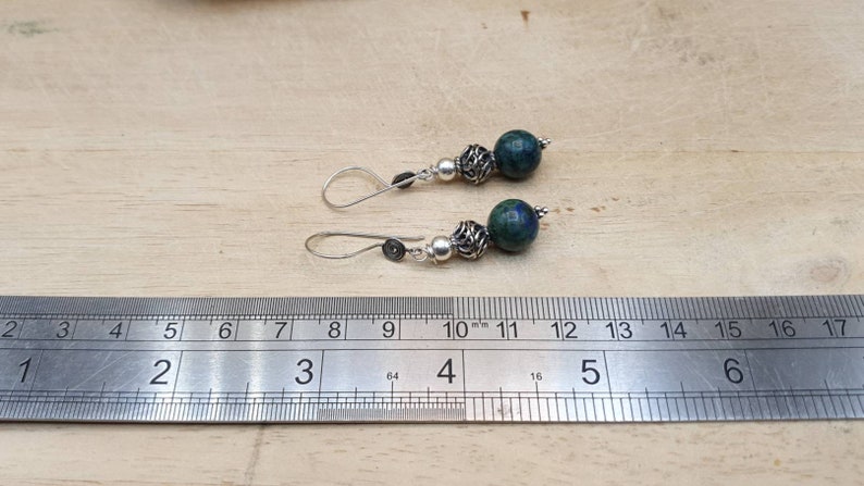 May include: A pair of handcrafted earrings featuring dark blue and green stone beads, silver-colored metal beads, and silver-colored ear wires. The earrings are displayed next to a ruler for size reference.