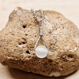 May include: A white gemstone pendant necklace with a silver chain. The pendant is a single round white stone with a silver wire wrap.